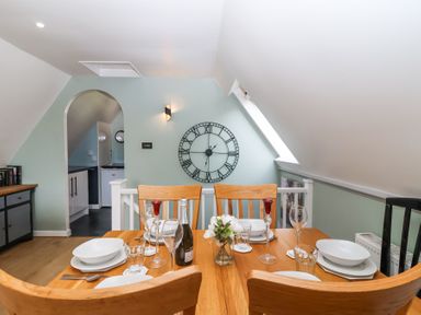 A dining area with a wooden table set with dishes glasses and a bottle under a wall clock at 23 Valley Lodge in Gunnislake