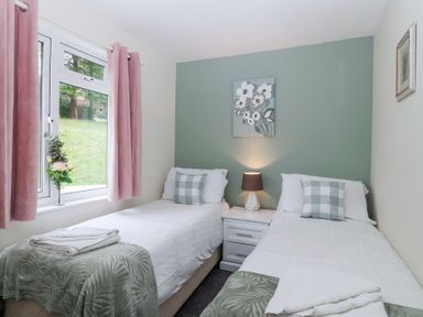 A bedroom with two single beds a nightstand with a lamp and pink curtains at 23 Valley Lodge in Gunnislake