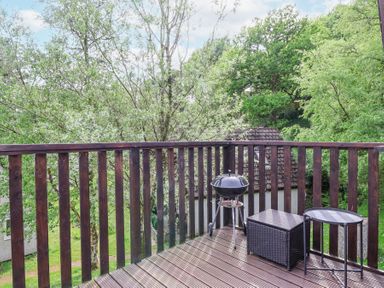 A wooden balcony with a charcoal grill a woven storage box and a small round table surrounded by trees at 23 Valley Lodge in Gunnislake
