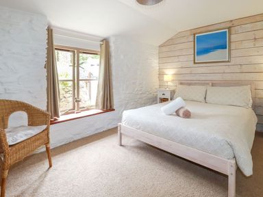 A bedroom with a bed a lamp on a nightstand a wicker chair and a window with curtains at Babes Cottage in Perranwell near Goonhavern