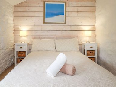 A bedroom with a bed with beige bedding and two rolled towels on it two bedside tables with lamps and baskets and a framed picture on a wooden paneled wall at Babes Cottage in Perranwell near Goonhavern