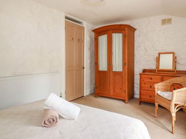 A bedroom with a bed and rolled towels a wooden wardrobe a wooden dresser with a mirror and a wicker chair at Babes Cottage in Perranwell near Goonhavern