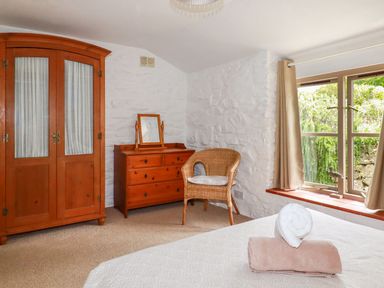 A bedroom with wooden wardrobe wooden dresser wicker chair and window with curtains at Babes Cottage in Perranwell near Goonhavern