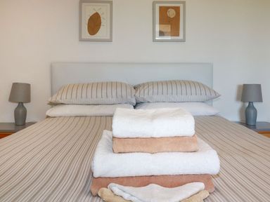 A bedroom with a bed with striped linens and stacked towels in the center with lamps and framed pictures on the wall at The Lookout in Perranwell near Goonhavern