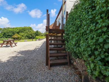 An outdoor area with wooden stairs next to a green hedge and a picnic table on a gravel surface at The Lookout in Perranwell near Goonhavern