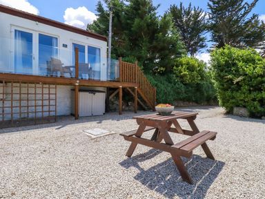 An outdoor gravel area with a wooden picnic table and a raised deck with chairs at The Lookout in Perranwell near Goonhavern