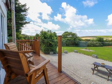 A wooden deck with two chairs and glass railing overlooking a gravel area and green field at The Lookout in Perranwell near Goonhavern