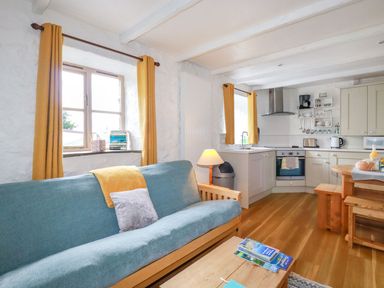 A living room with a blue sofa and yellow curtains next to a kitchen with wooden dining furniture at Hollies Cottage in Perranwell near Goonhavern