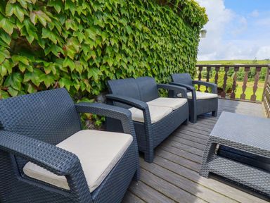 An outdoor wooden deck with dark wicker chairs and a table next to a wall covered in green ivy at Hollies Cottage in Perranwell near Goonhavern