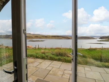 A view of a grass lawn and water with hills in the distance seen through open glass doors at Insh Cullipool Isle Of Luing