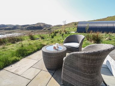 Two wicker chairs and a wicker table with two mugs on a stone patio overlooking grassy hills and a corrugated metal structure at Sula Cullipool Isle Of Luing