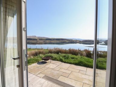 View through open glass doors onto a stone patio with grass and a body of water with hills in the background at Scarba in Cullipool Isle Of Luing