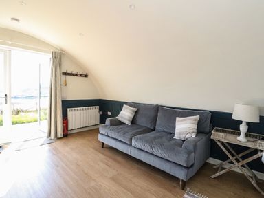 A living room with a gray sofa two pillows a wooden side table with a lamp a curtain a coat rack and a glass door at Scarba in Cullipool Isle Of Luing