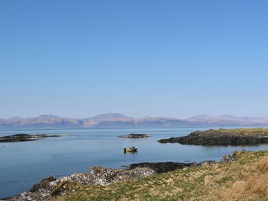 A small boat on calm water near rocky shoreline with mountains in the background at Torsa Cullipool Isle Of Luing
