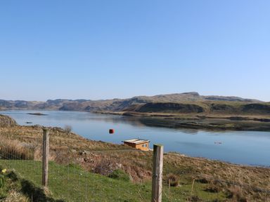 A coastal landscape with a small wooden structure near calm water and hills in the background at Torsa Cullipool Isle Of Luing