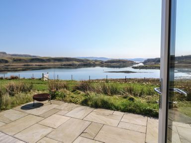 A view of a tiled patio with a fire pit and grassy area overlooking a body of water and hills at Torsa Cullipool Isle Of Luing