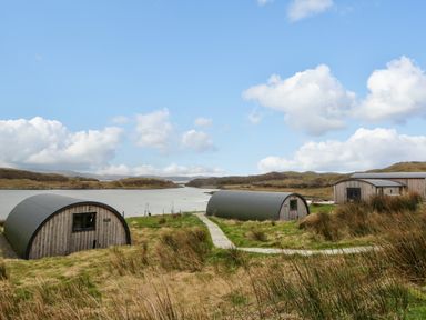 Two semi cylindrical buildings with wood siding near water with hills in background at Fladda Cullipool Isle Of Luing