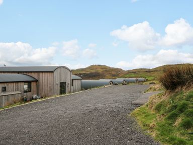 A gravel road leading to wooden buildings with curved roofs next to grassy hills under a blue sky at Fladda in Cullipool Isle Of Luing