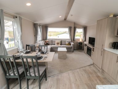 A living area with a dining table set with plates and glasses a sofa with cushions a coffee table a television on a cabinet and windows with curtains at Meadow View in West Thirston Felton