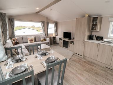 An open plan living room and kitchen with a dining table set for four a grey sofa with cushions a TV on a cabinet and kitchen cabinets with appliances at Meadow View in West Thirston Felton