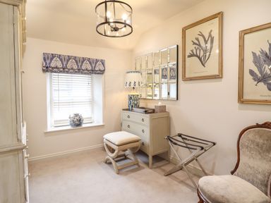 A room with a window with blinds and a blue patterned valance a chest of drawers with a lamp and mirror and framed botanical prints at 6 Uppergate Street Rose Cottage in Conwy