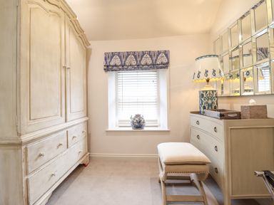 A room with a large wardrobe a window with blinds and valance a dresser with a lamp and mirror and a cushioned bench at 6 Uppergate Street Rose Cottage in Conwy