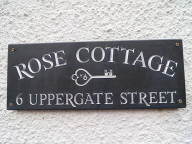 A black address plaque with white engraved text reading Rose Cottage 6 Uppergate Street on a textured white wall at 6 Uppergate Street Rose Cottage Conwy
