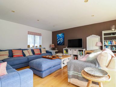 A living room with blue sofas a large ottoman a wooden coffee table a television on a white storage unit and bookshelves at Goatsland in Trebetherick