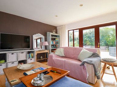A living room with a pink covered sofa a wooden coffee table a large TV and a bookshelf at Goatsland in Trebetherick