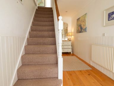 A carpeted staircase beside a hallway with a wooden floor a rug a radiator a dresser with a lamp and wall art at Goatsland in Trebetherick