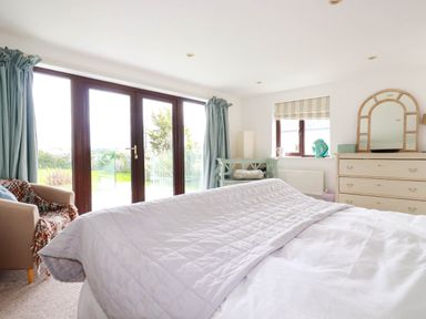 A bedroom with a bed covered with white bedding a dresser with a mirror a window and glass doors with blue curtains at Goatsland in Trebetherick