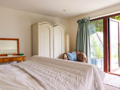 A bedroom with a bed, white wardrobe, wooden vanity with mirror, armchair with cushion, and open glass door at Goatsland in Trebetherick