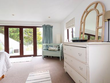 A room with a white dresser a large mirror a blue bench and large glass doors at Goatsland in Trebetherick