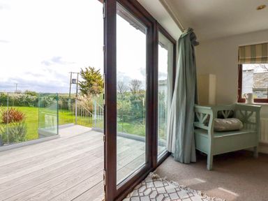 A room with a green wooden bench pillow and curtains opening to a balcony with glass railing and garden at Goatsland in Trebetherick