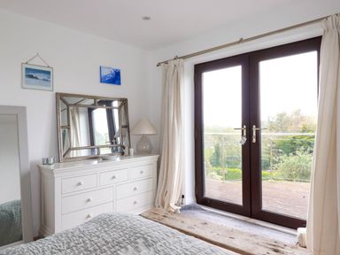 A bedroom with a white dresser mirror table lamp and glass doors opening to a balcony at Goatsland in Trebetherick