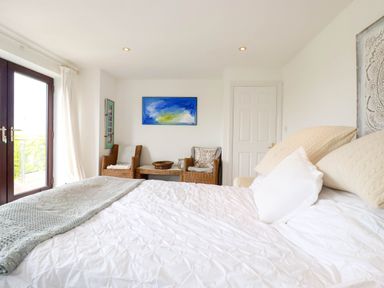 A bedroom with a bed white bedding two armchairs and a table near a door and glass patio doors at Goatsland in Trebetherick