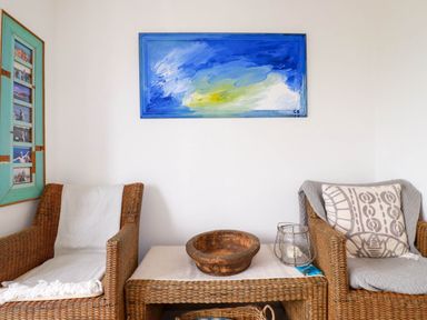A sitting area with two wicker chairs a wicker table with a bowl and lantern a blue painting on the wall at Goatsland in Trebetherick
