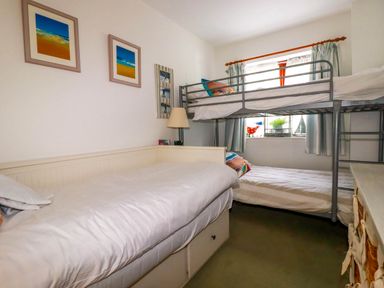 A small bedroom with a bunk bed and a single bed with a lamp and framed pictures on the wall at Goatsland in Trebetherick