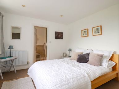A bedroom with a bed with pillows and white bedding a side table with a lamp and an open door leading to a bathroom at Goatsland in Trebetherick
