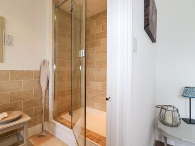 A bathroom corner with a glass shower door and beige tiled walls next to a hallway with a table holding a lamp and a decorative candle holder at Goatsland in Trebetherick