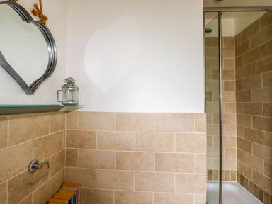 A bathroom corner with beige tiled walls a heart-shaped mirror a small glass shelf a lantern and a shower area at Goatsland in Trebetherick