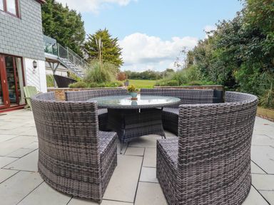 An outdoor patio area with a round glass table surrounded by curved wicker chairs and green plants at Goatsland in Trebetherick