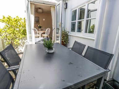 An outdoor patio with a black table and four black chairs with potted plants and an open door leading to a dining room with a round table and white chairs at Shrimp Cottage Brewers Quay Harbour