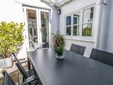 An outdoor patio with a black table and six chairs a small plant on the table potted plants near a white wall and glass doors at Shrimp Cottage in Brewers Quay Harbour