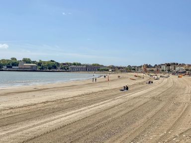 A sandy beach with people sitting and walking near the water and buildings in the background at Shrimp Cottage in Brewers Quay Harbour