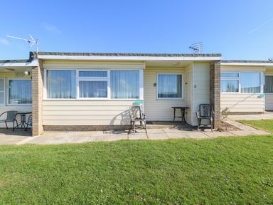 A single story building with windows chairs and tables on a stone patio in front of a lawn at Chalet 37 in Hemsby