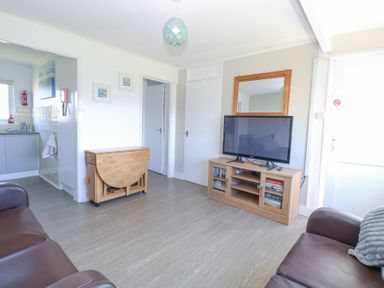 A living room with two brown sofas a wooden tv cabinet with a tv and books a folding wooden table and a kitchen visible through a doorway at Chalet 37 in Hemsby