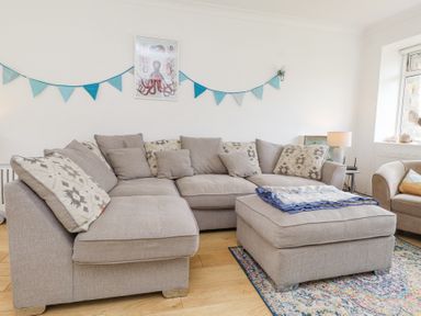 A living room with a beige sectional sofa, matching ottoman, blue bunting on wall, and an octopus picture at 3 Glyn Y Mor in Llanbedrog