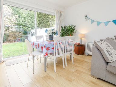 A dining area with a table covered in a lobster-patterned tablecloth surrounded by four chairs next to a large window and a gray sofa with patterned cushions at 3 Glyn Y Mor in Llanbedrog