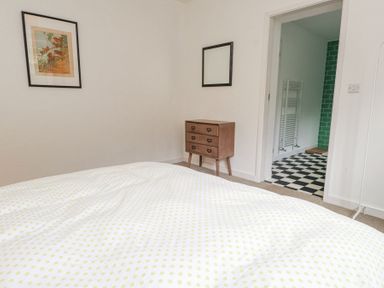 A bedroom with a bedspread with yellow dots a wooden dresser a framed picture and an open door to a bathroom with black and white checkered flooring at 3 Glyn Y Mor in Llanbedrog
